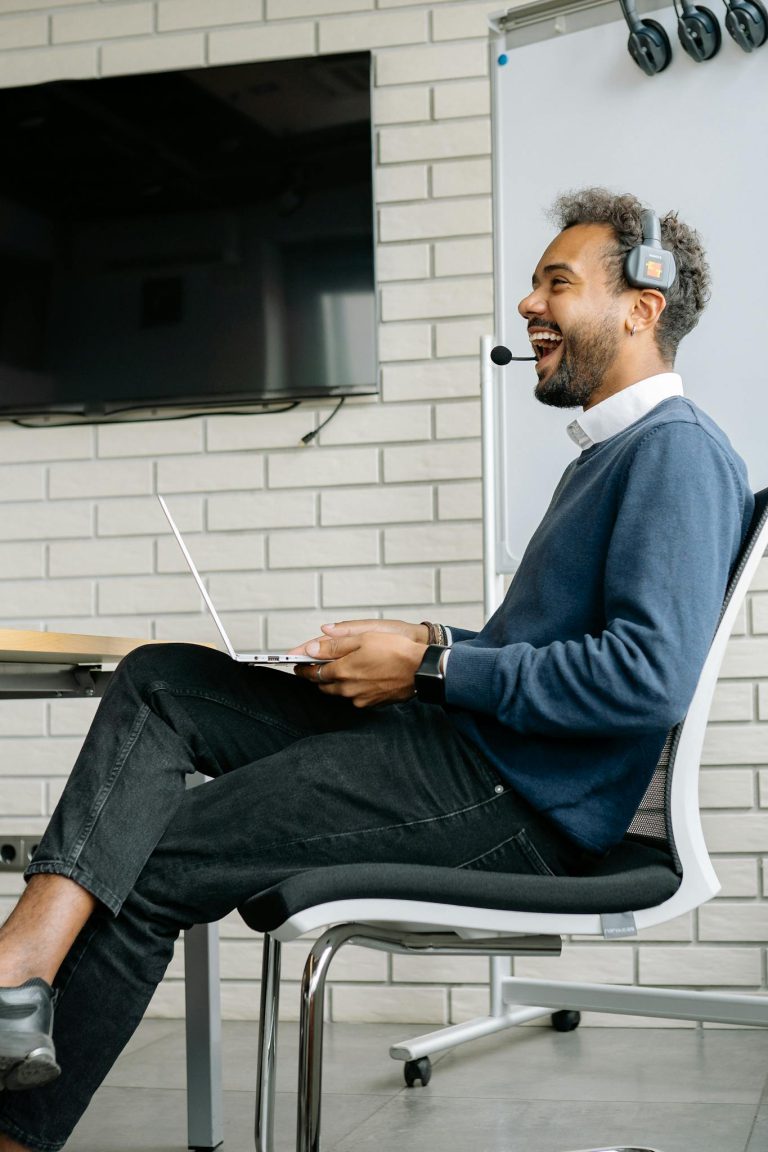 Friendly customer service agent laughing while working on his laptop with a headset in a modern office.